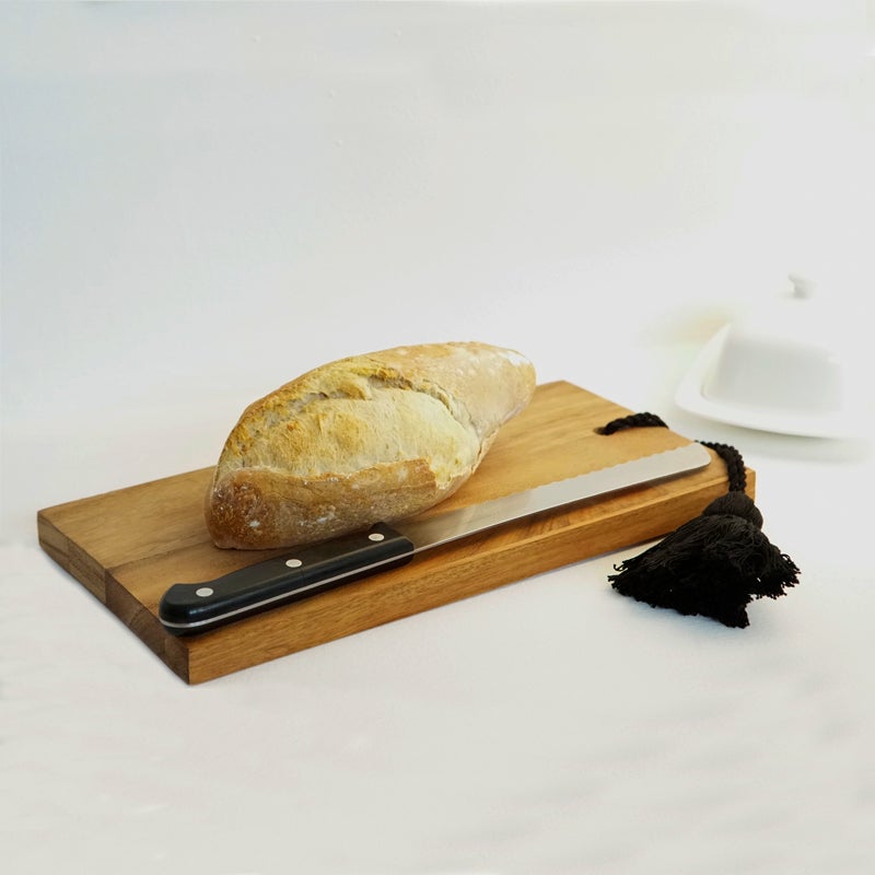 Una tabla para cortar hecha de madera con un pan y un cuchillo sobre ella decorada con una borla de hilo negro